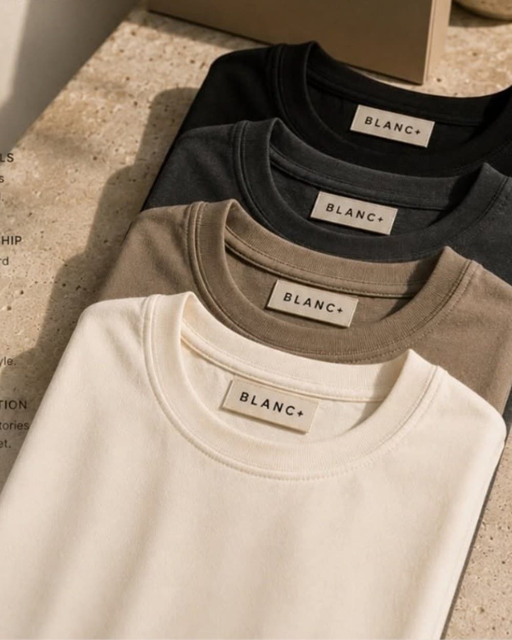 Four Blanc+ tees folded in cream, taupe, charcoal and black beside a kraft gift box